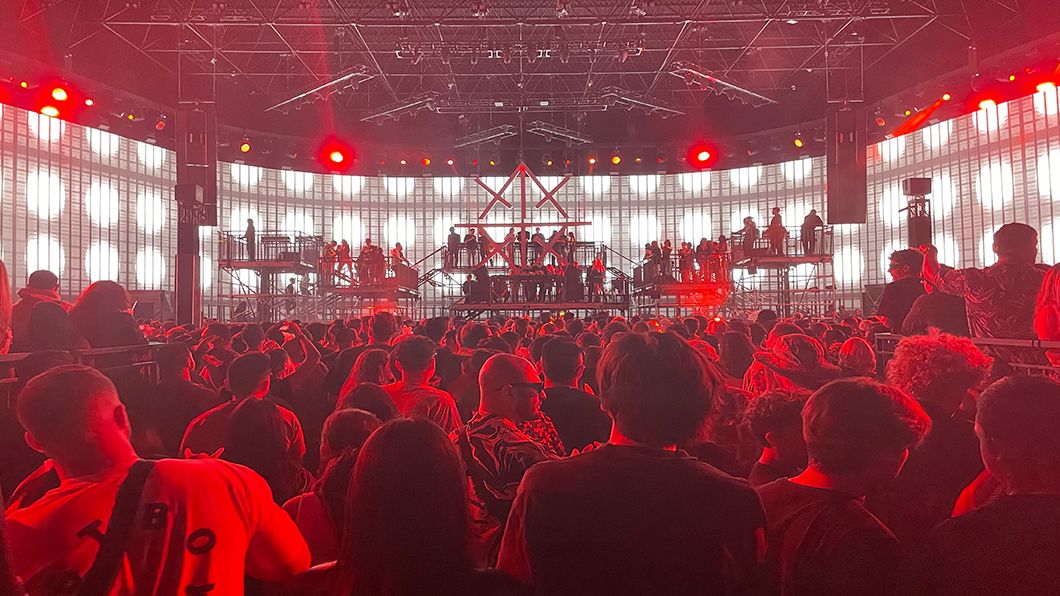 An in-the-crowd perspective looking towards the stage, where red stage lights cast an intense glow over a large group of concertgoers.