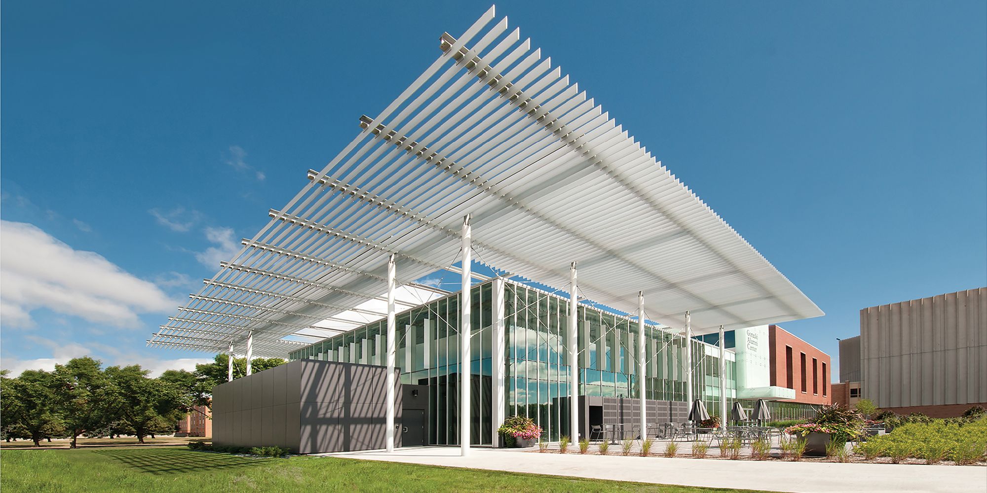 Modern building with a white slatted roof, glass walls, and outdoor seating under a bright blue sky.