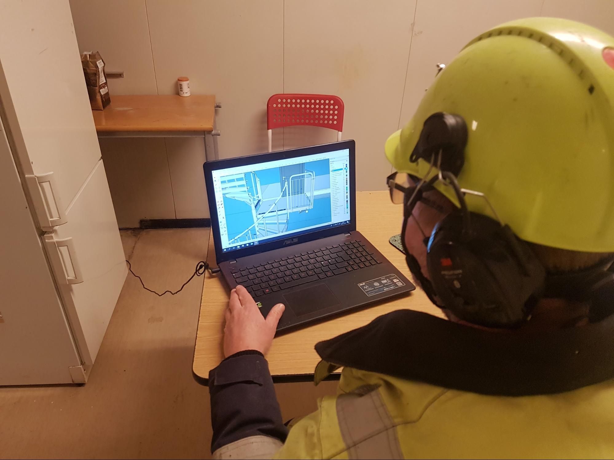 Worker in hard hat and safety vest reviews architectural plans on a laptop.