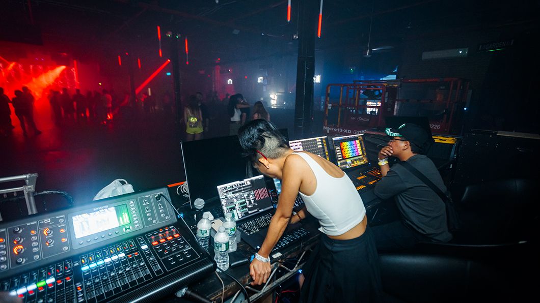 Sayoko Osada, wearing a white tank top, adjusts controls at a concert venue, with the audience gathering in the background under red stage lighting